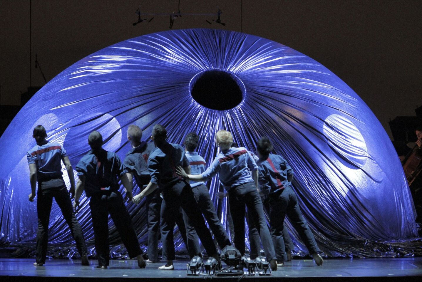 A group of male dancers tilt to their left as the circular centerpiece is covered in a plum colored fabric.