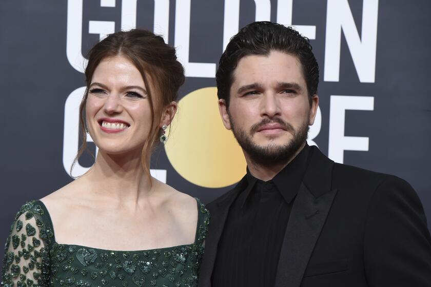 Rose Leslie and Kit Harington pose and smile together in formal attire against a black background with white lettering.