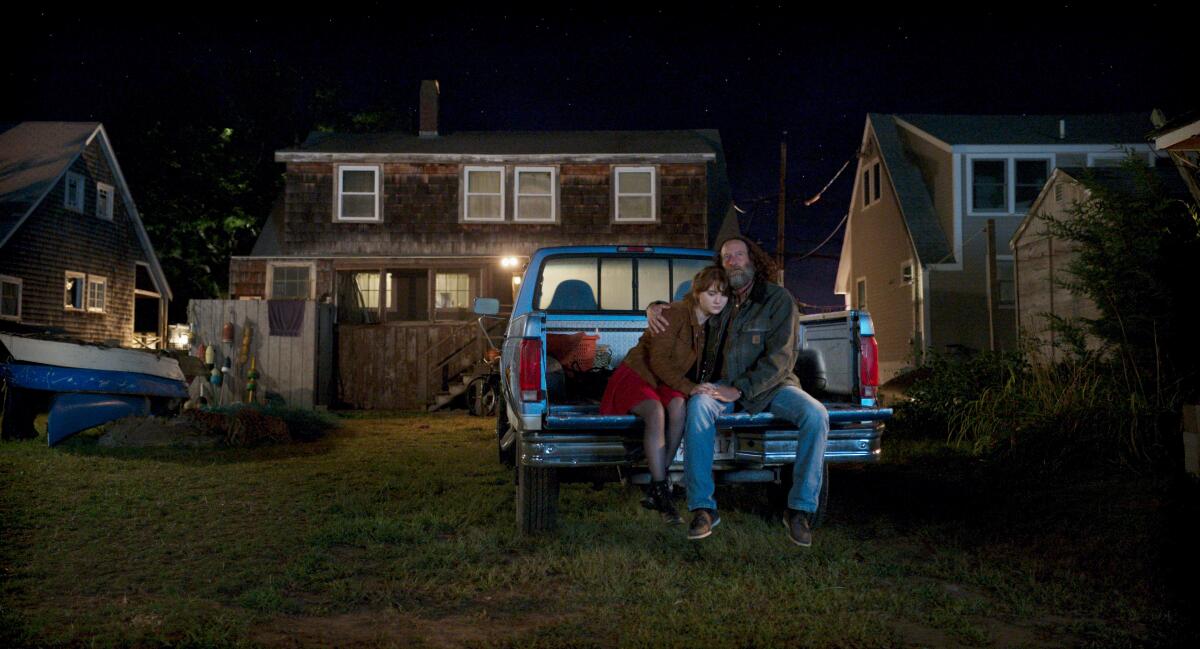 A father with his arm around his daughter in the bed of a pick-up truck outside a house in the dark