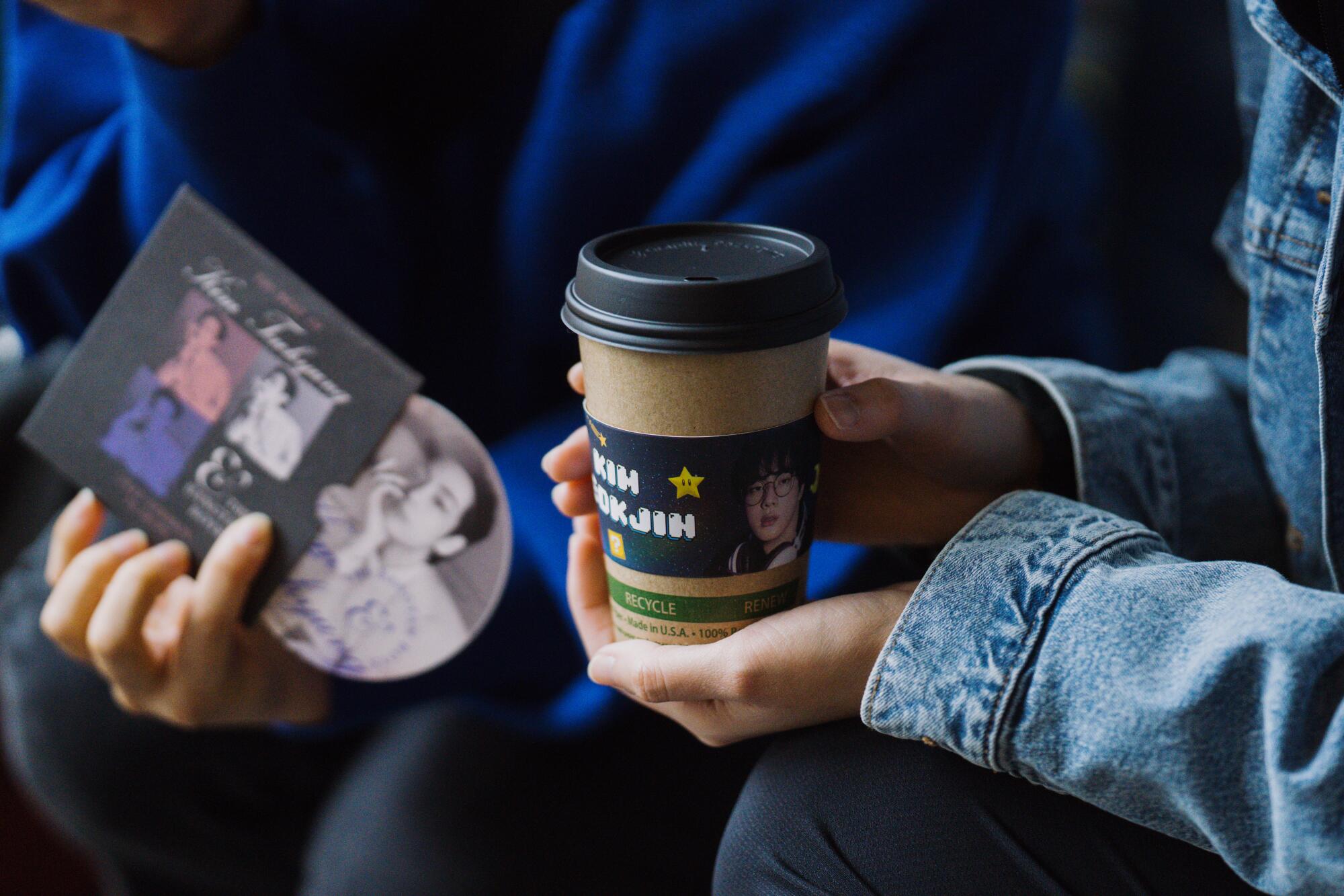 Hands holding a K-pop CD and a drink with a cupsleeve