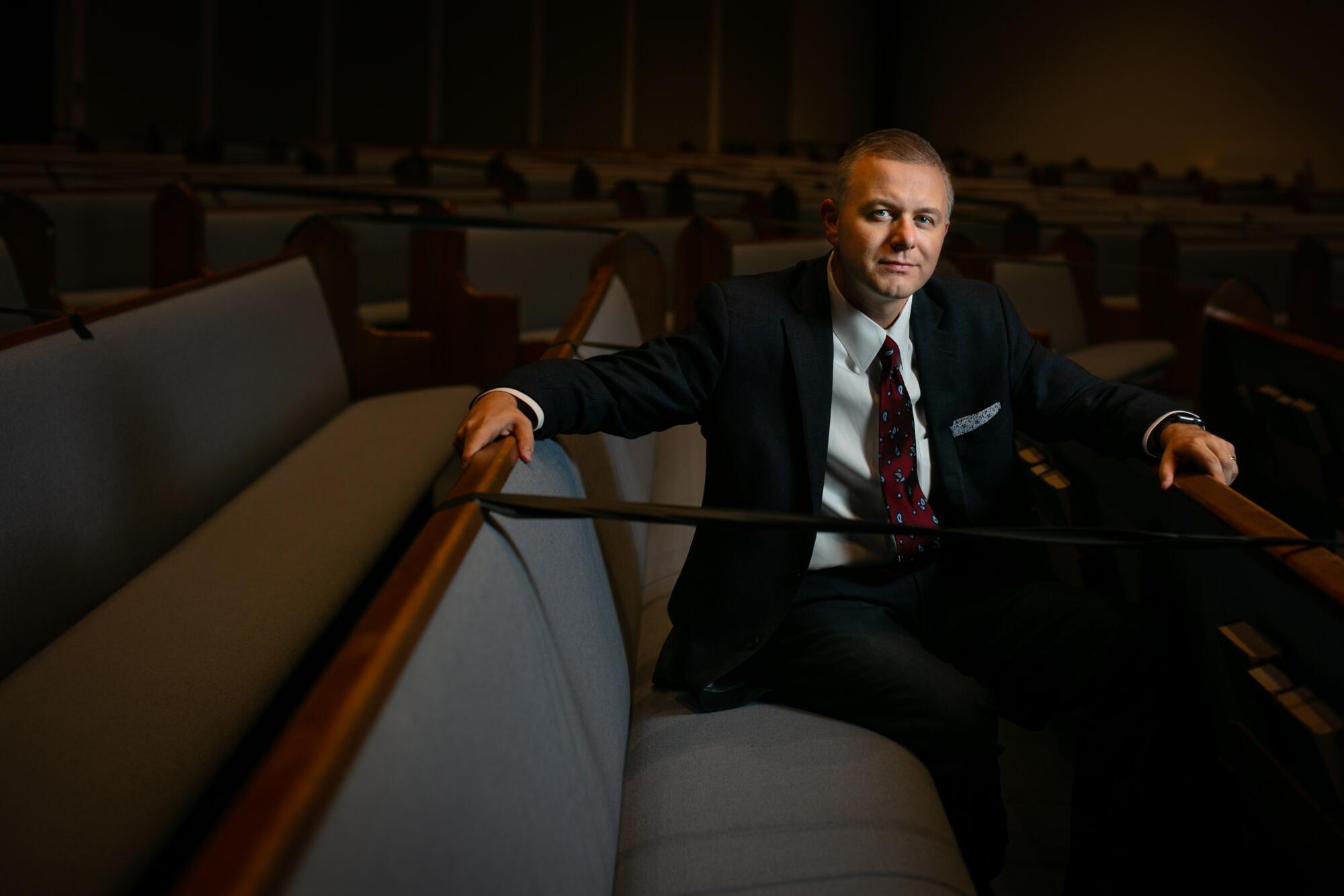 A man in a suit sits for a portrait on a padded church pew.