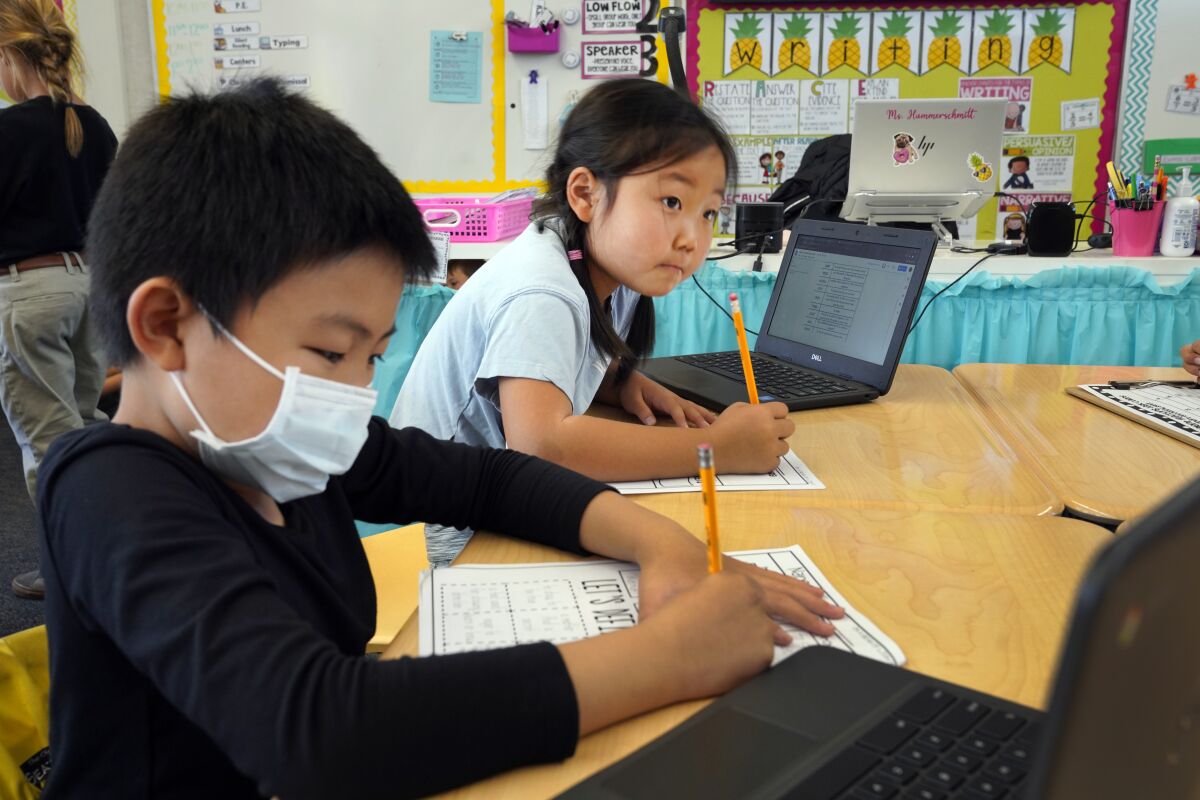Two 8-year-olds, one with a mask, one without, work at their desks. 