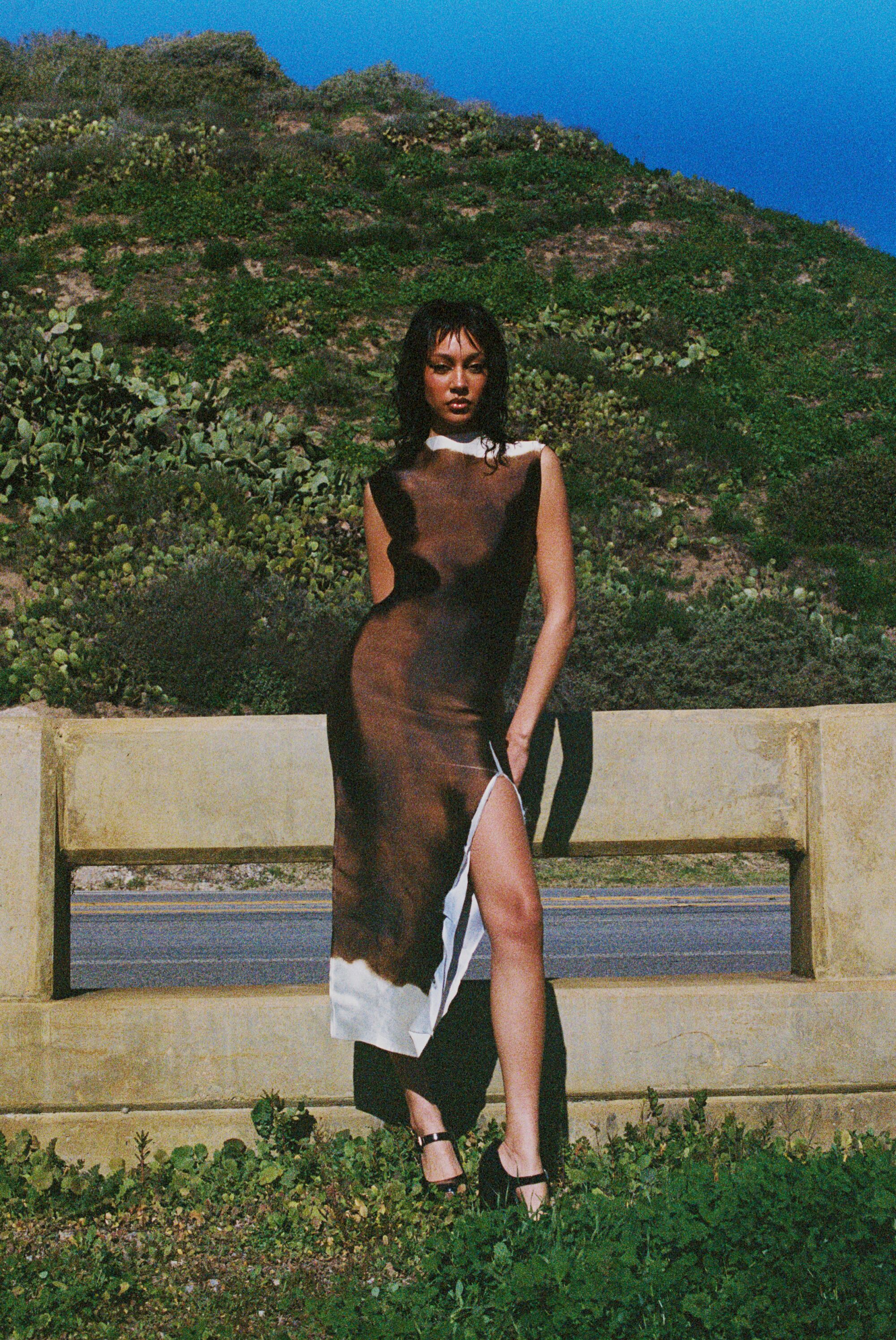 The detox for L.A. life you deserve is an all-black wardrobe Model wears a black dress in the sunshine, a green hill behind her