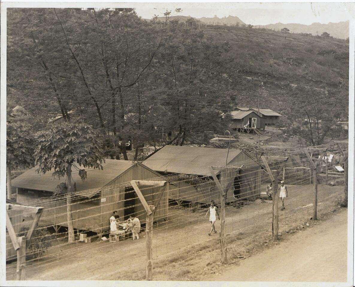 A young man walks along a barbed wire fence as other detainees gather outside barracks at Honouliuli internment camp on Oahu during World War II.