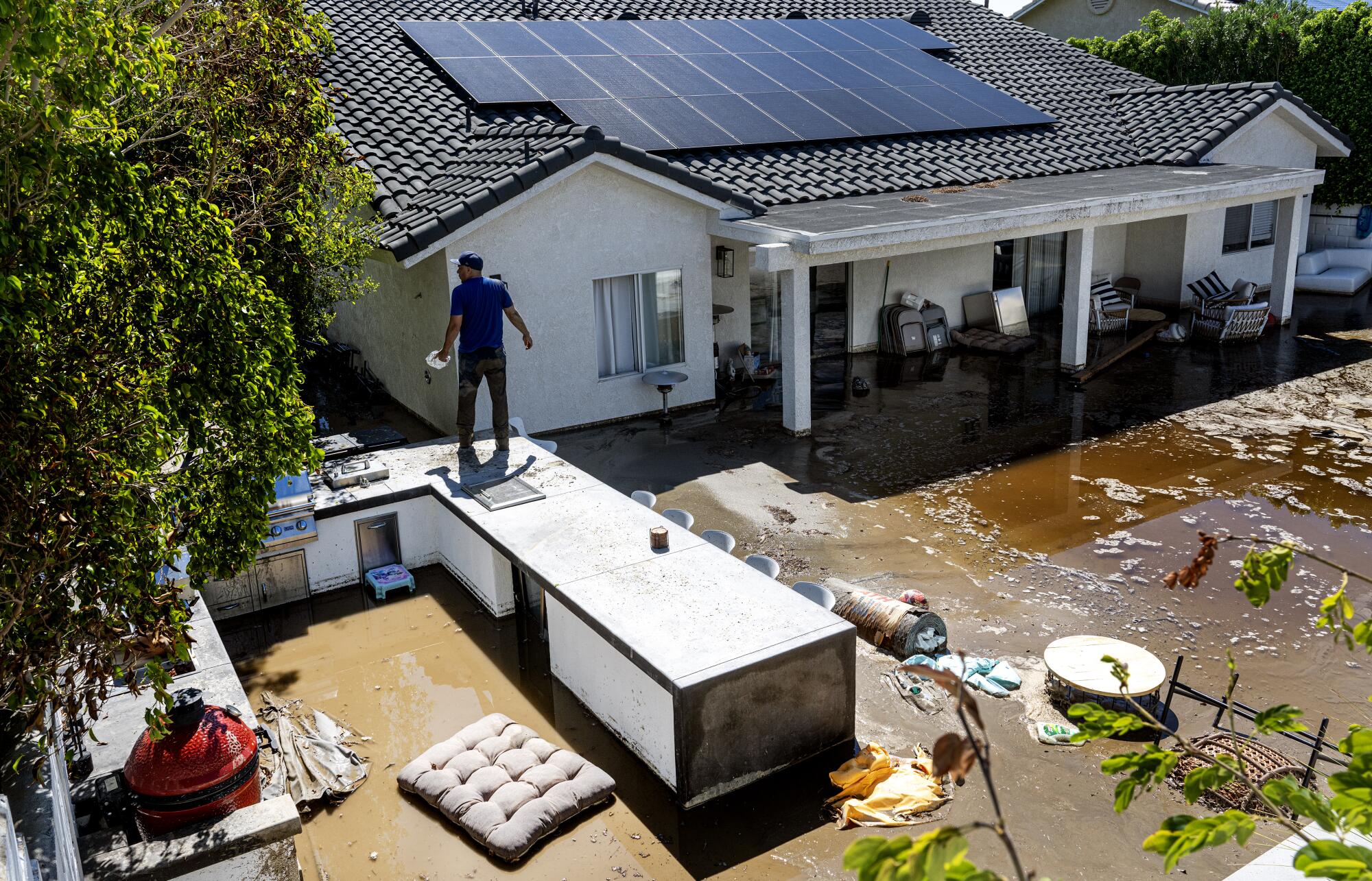 Un homme se tient au sommet d’un barbecue extérieur dans une maison inondée.