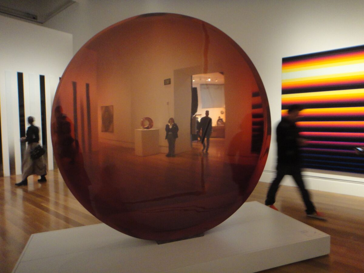 A security guard is visible through a transparent resin sculpture in red by De Wain Valentine in a gallery