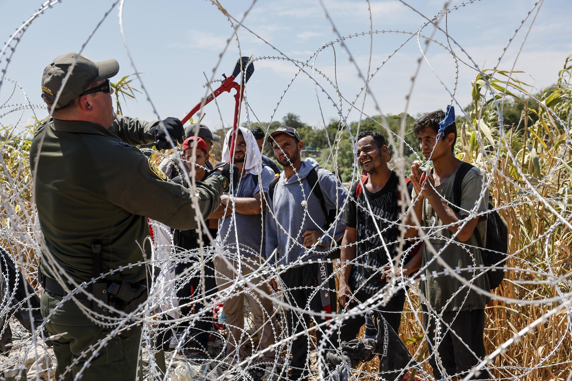 A Border Patrol agent uses wire cutters on coils of razor wire as people wait.