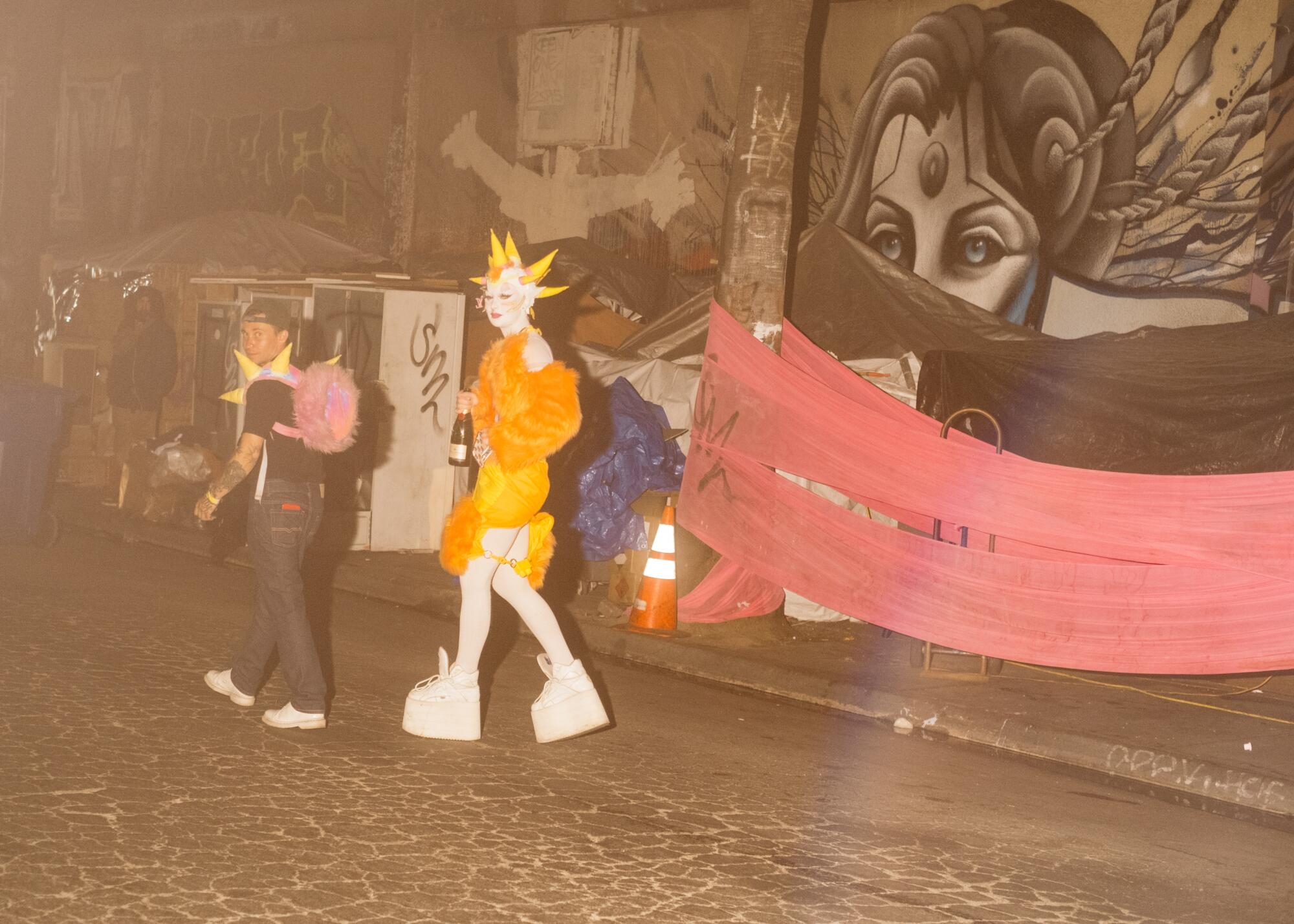 People in extravagant outfits walk outside a muraled warehouse on a street at night in LA.