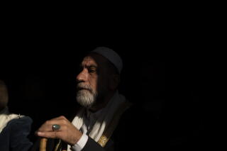 A man with a gray beard and white skullcap is shown at a meeting