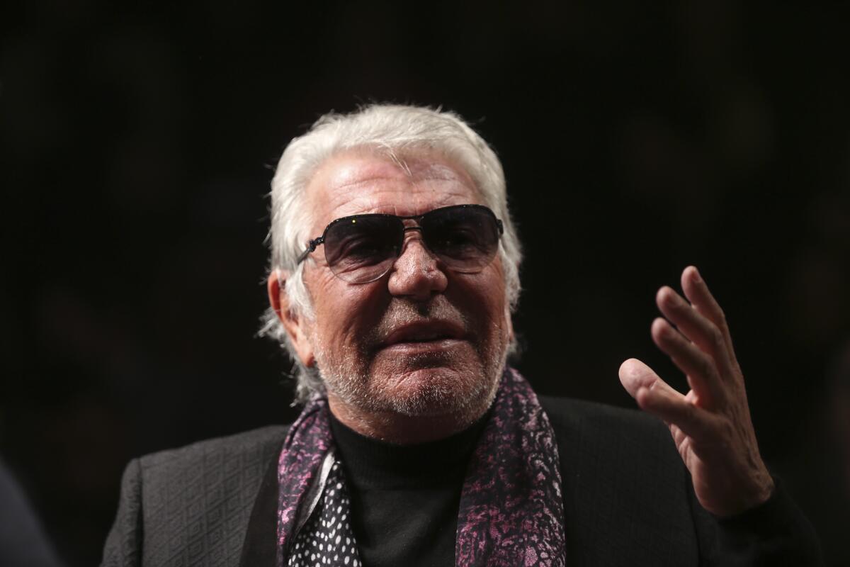 An older man in sunglasses, a black jacket and purple scarf with a thick head of white hair gestures with one hand
