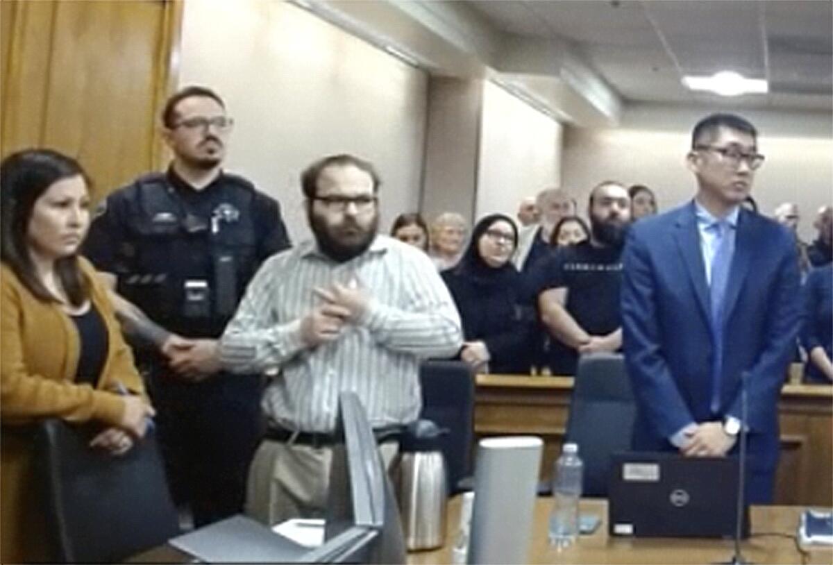 A bearded man wearing glasses, third from left, stands while flanked by other people in a courtroom