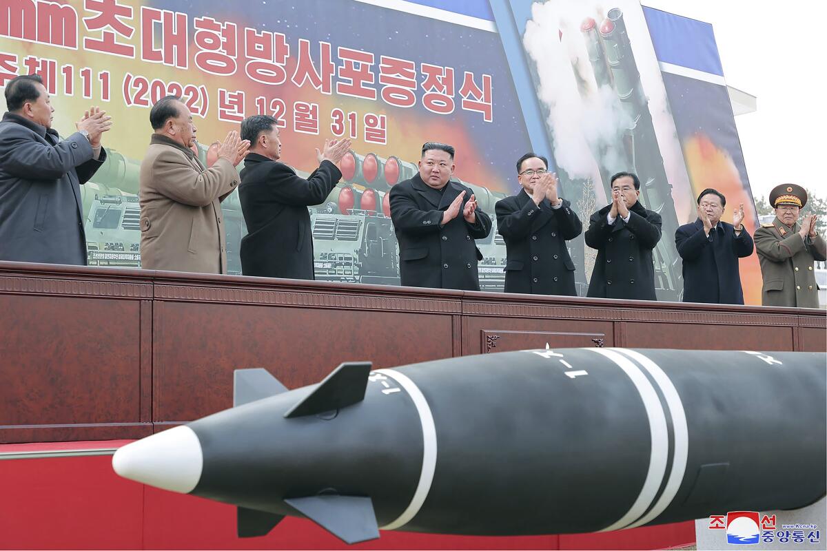 Men clap on a stage in front of a Korean-language poster showing missiles, as a missile lies in the foreground. 