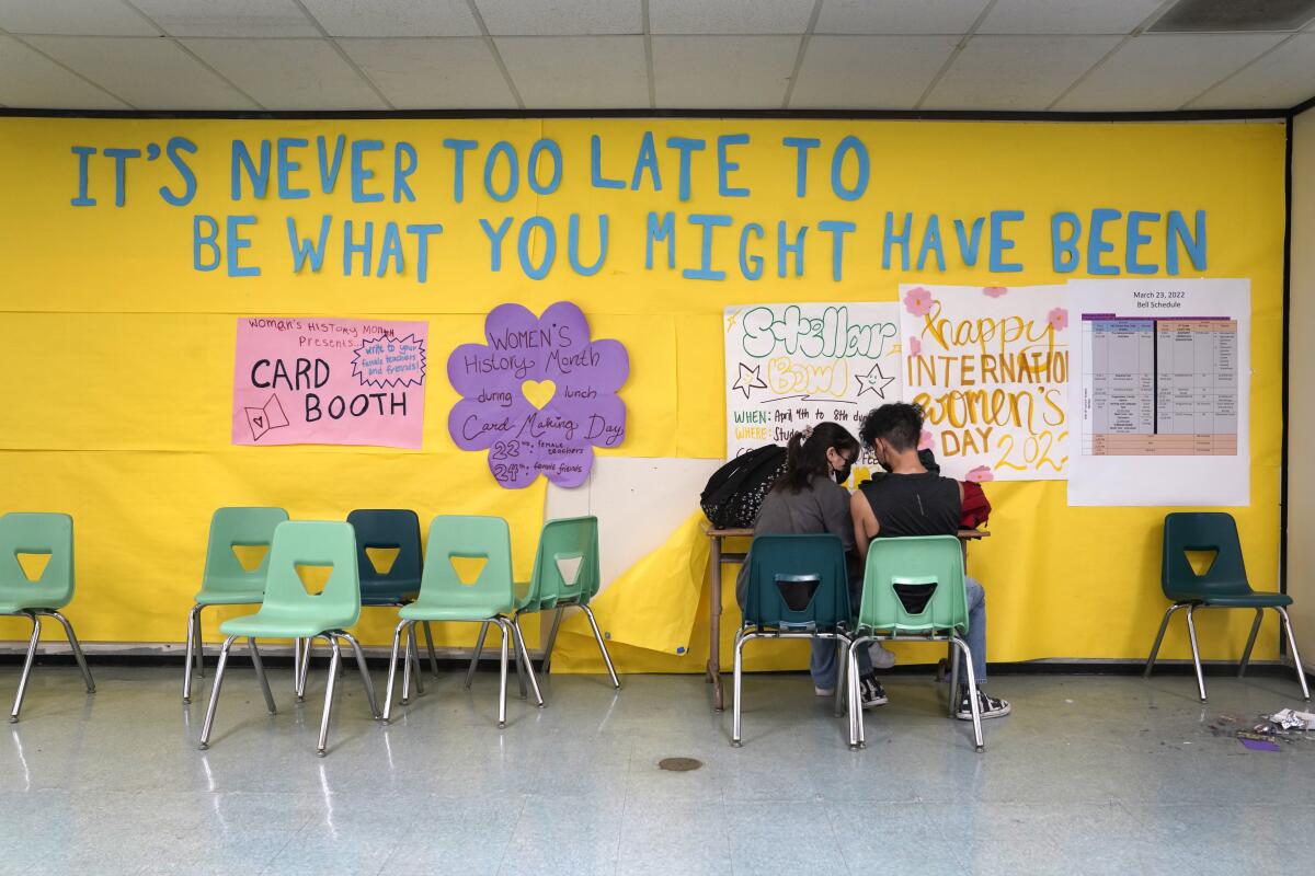 Two students huddle in a classroom with motivational signs on the wall
