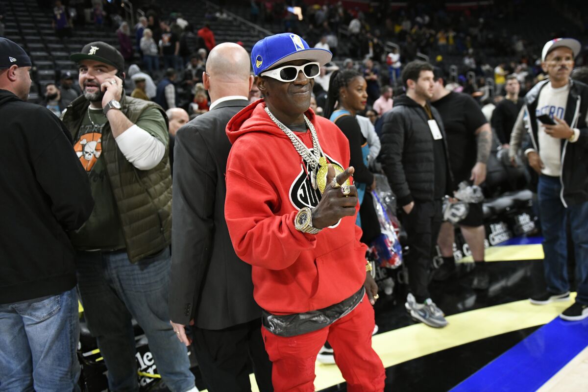 A man wearing a large clock around his neck, sunglasses and a ballcap stands in front of a crowd on a gym floor