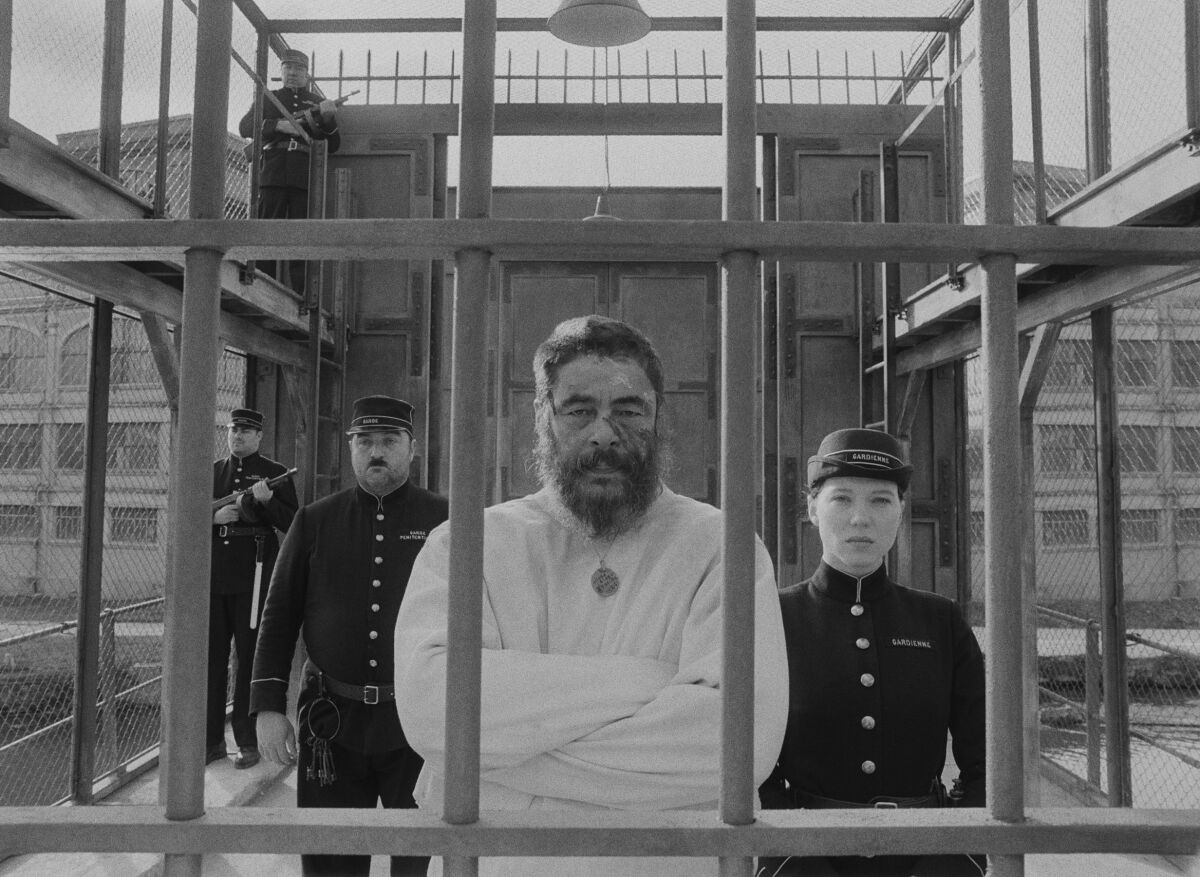A male guard and a female guard flank a straitjacketed prisoner staring out through prison bars.