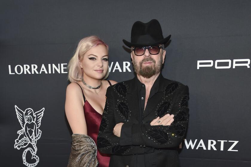 Kaya Stewart in a burgundy gown standing behind Dave Stewart in a black hat, sunglasses and a black suit