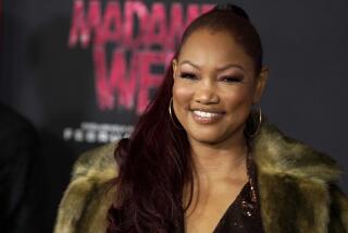 Garcelle Beauvais wearing a fur coat and hoop earrings smiling and posing against a black background with red text