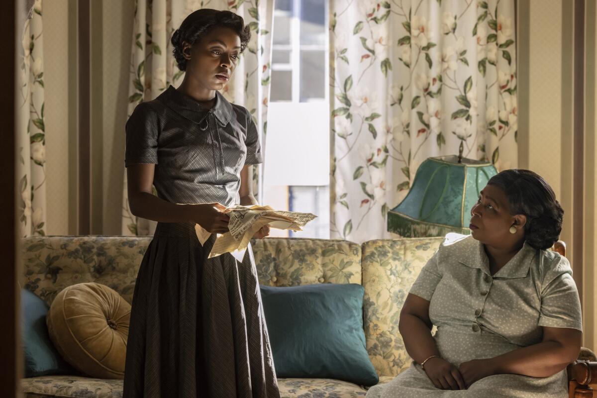 A standing woman, holding papers, talks to a seated woman in a living room.