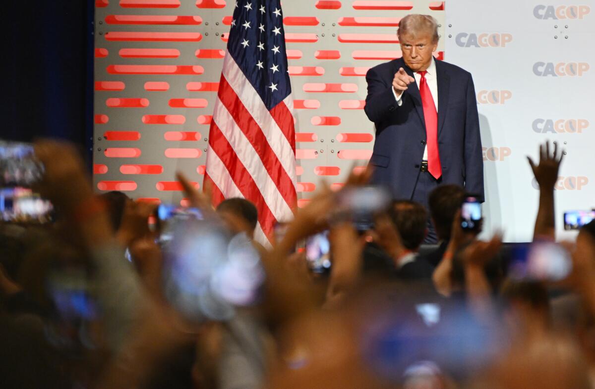 Donald Trump standing on a stage with a flag and pointing into the audience