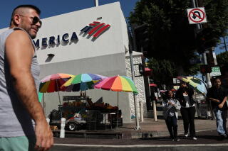 A man stands outside a bank, with colorful umbrellas providing shade near other pedestrians
