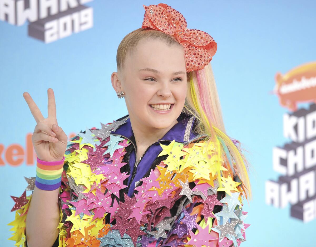 A young woman in a multicolored outfit and big hair bow holds up her fingers in a peace sign.