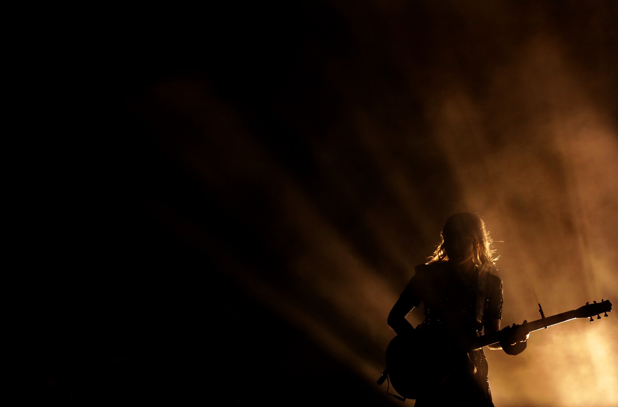 A woman is backlighted while she plays a guitar on a dark stage.
