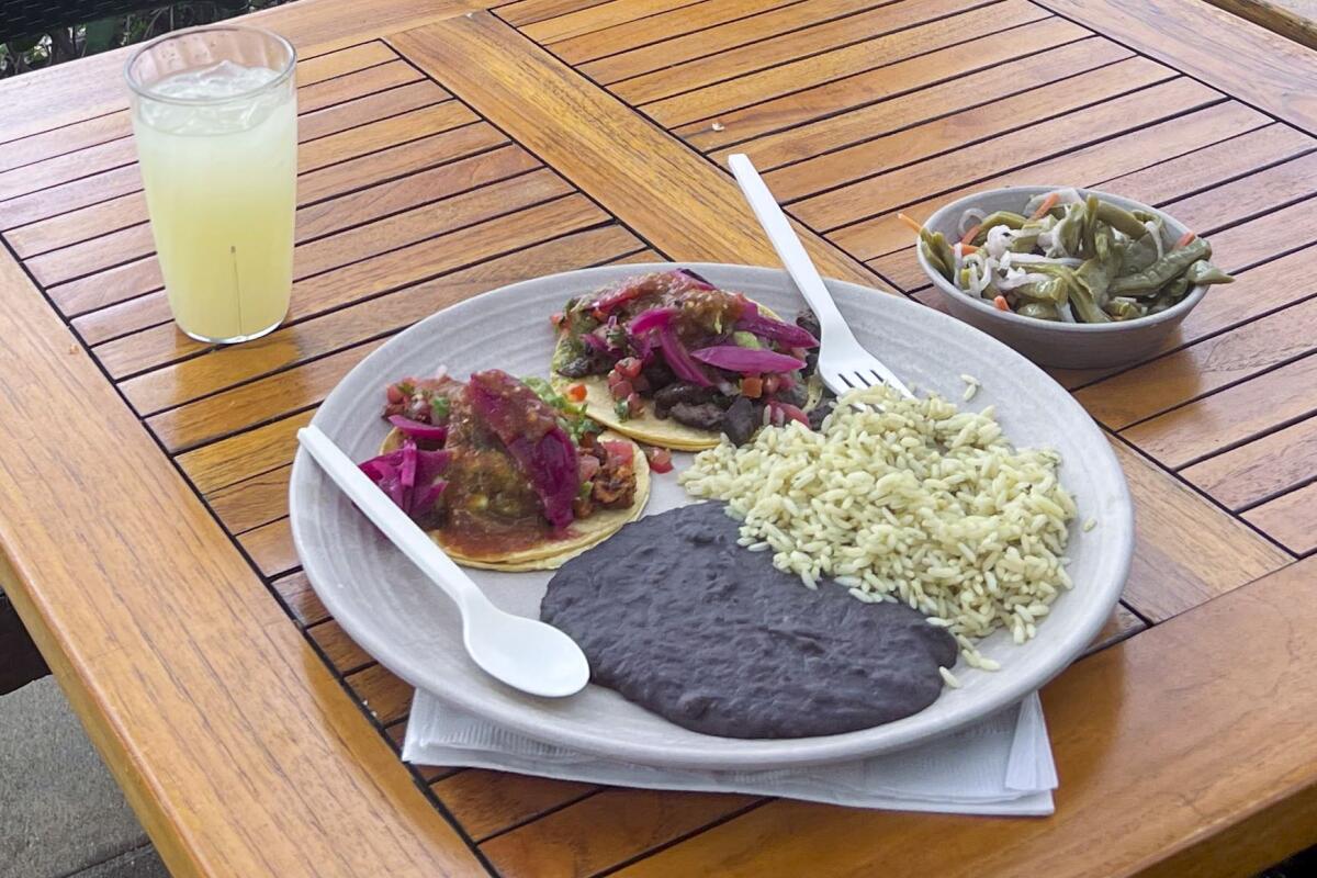 A plate with two tacos, rice and beans, with a side of nopales salad and lemonade, on a wooden table.