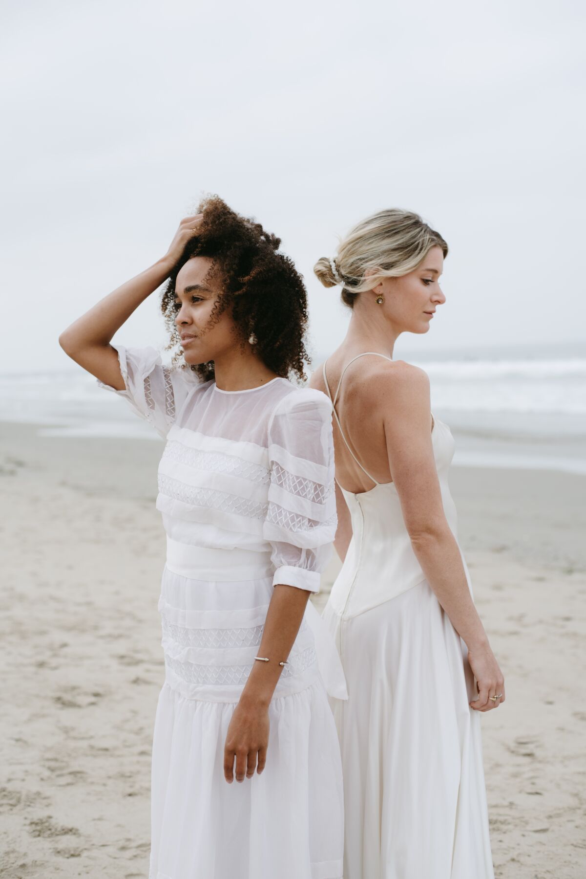 Two women in vintage bridal gowns stand back to back on a beach.