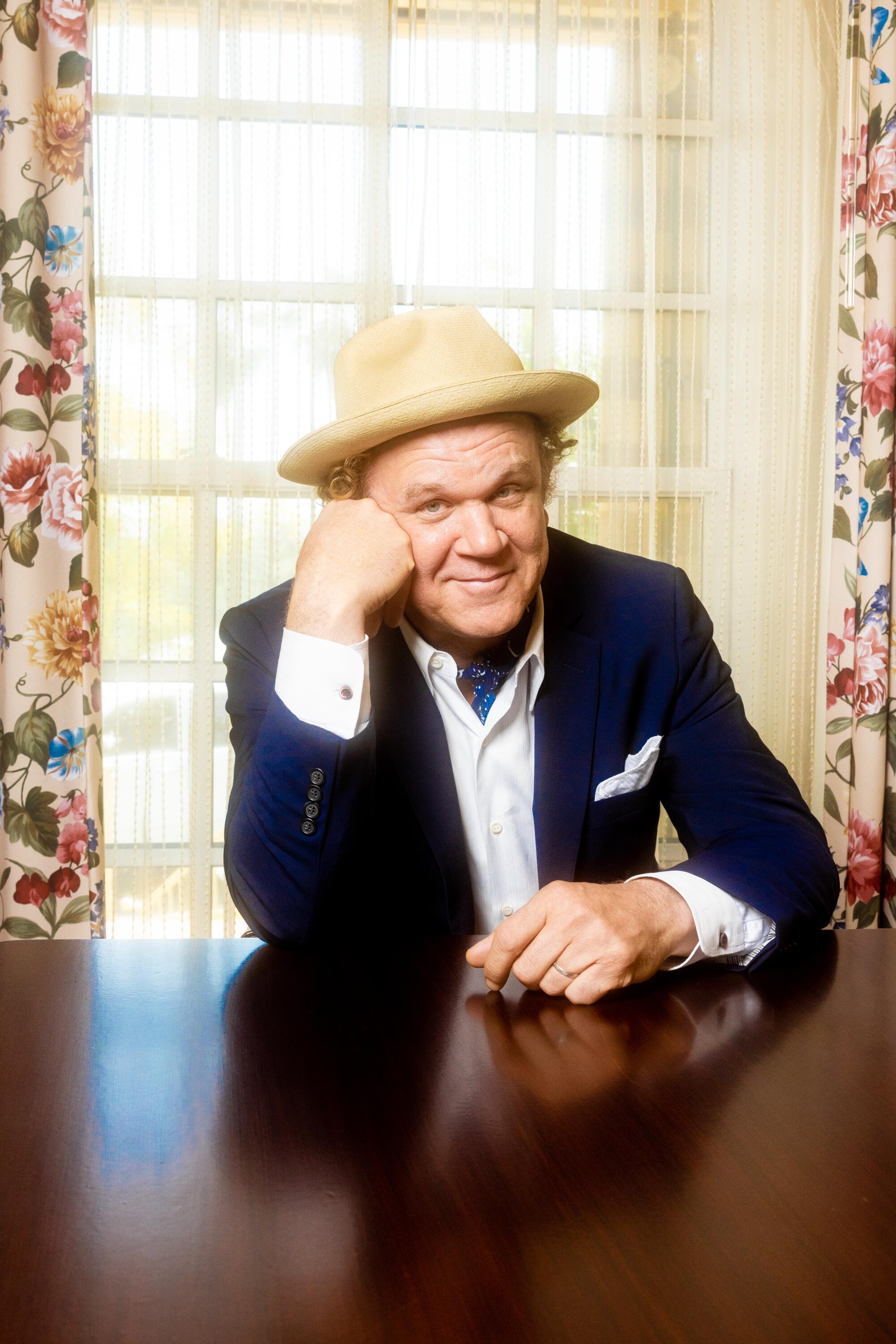 John C. Reilly leans on a wooden dining table.