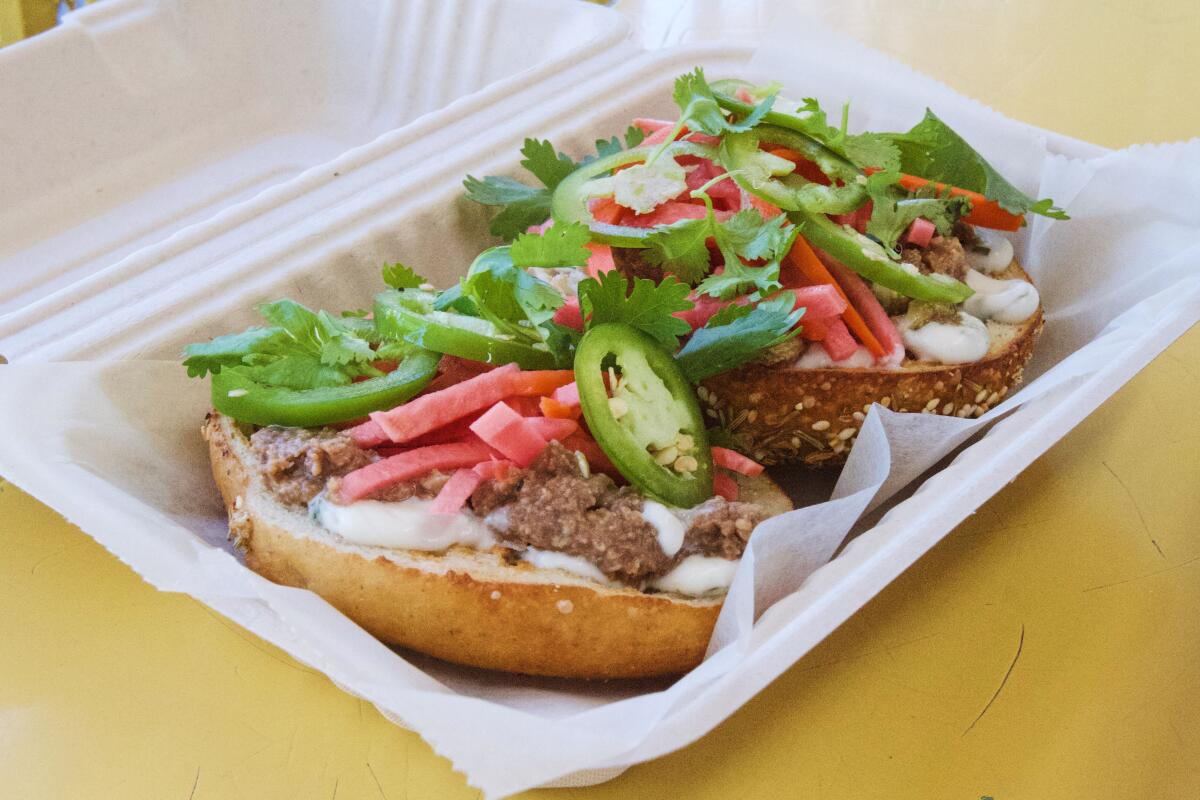 An open-faced bagel topped with walnut spread, jalapeos and pickled veg in a takeout box on A yellow bench