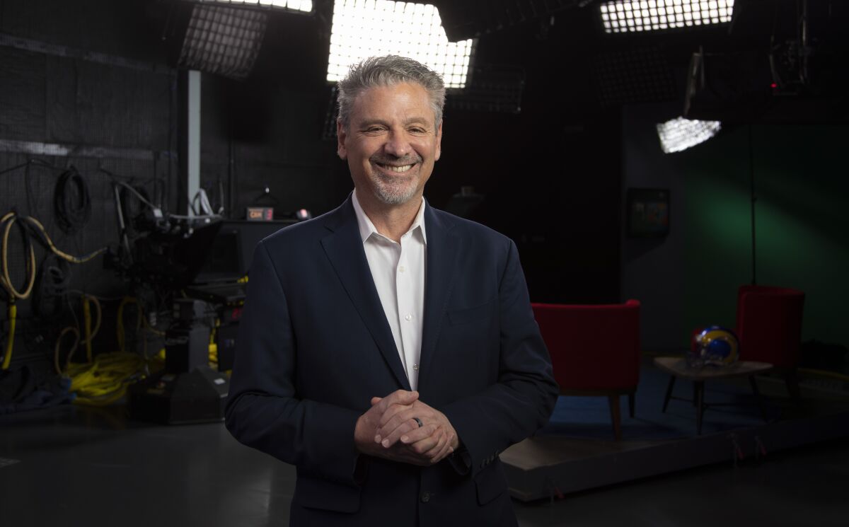 Fred Roggin stands inside a TV studio with lights behind him.