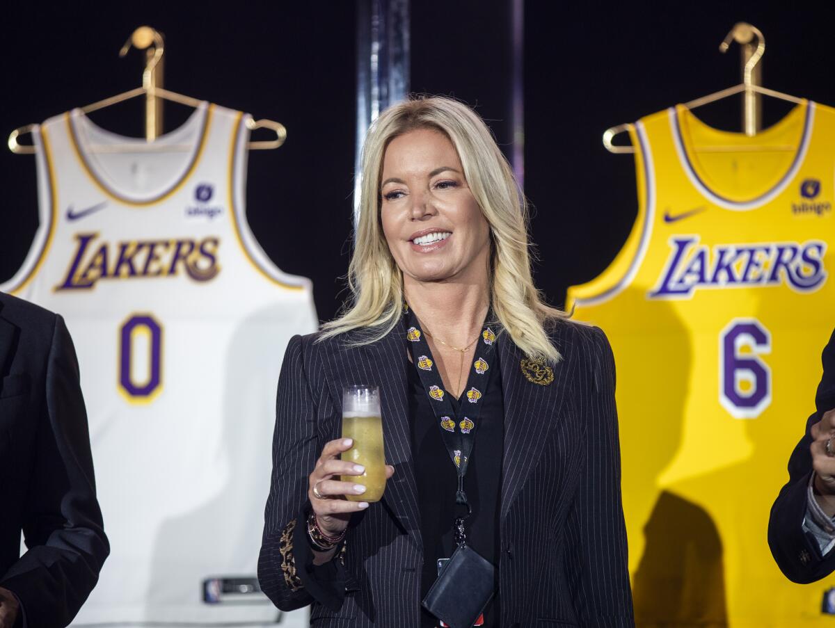 A blond woman holds a glass of liquid and stands before two Los Angeles Lakers jerseys, one white and one gold.