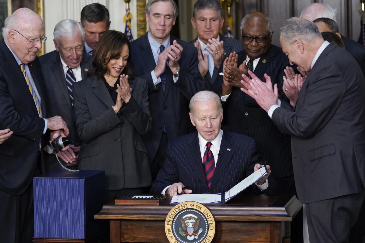 President Biden sits at a desk looking at documents as Vice President Harris and a group of lawmakers stand and applaud.