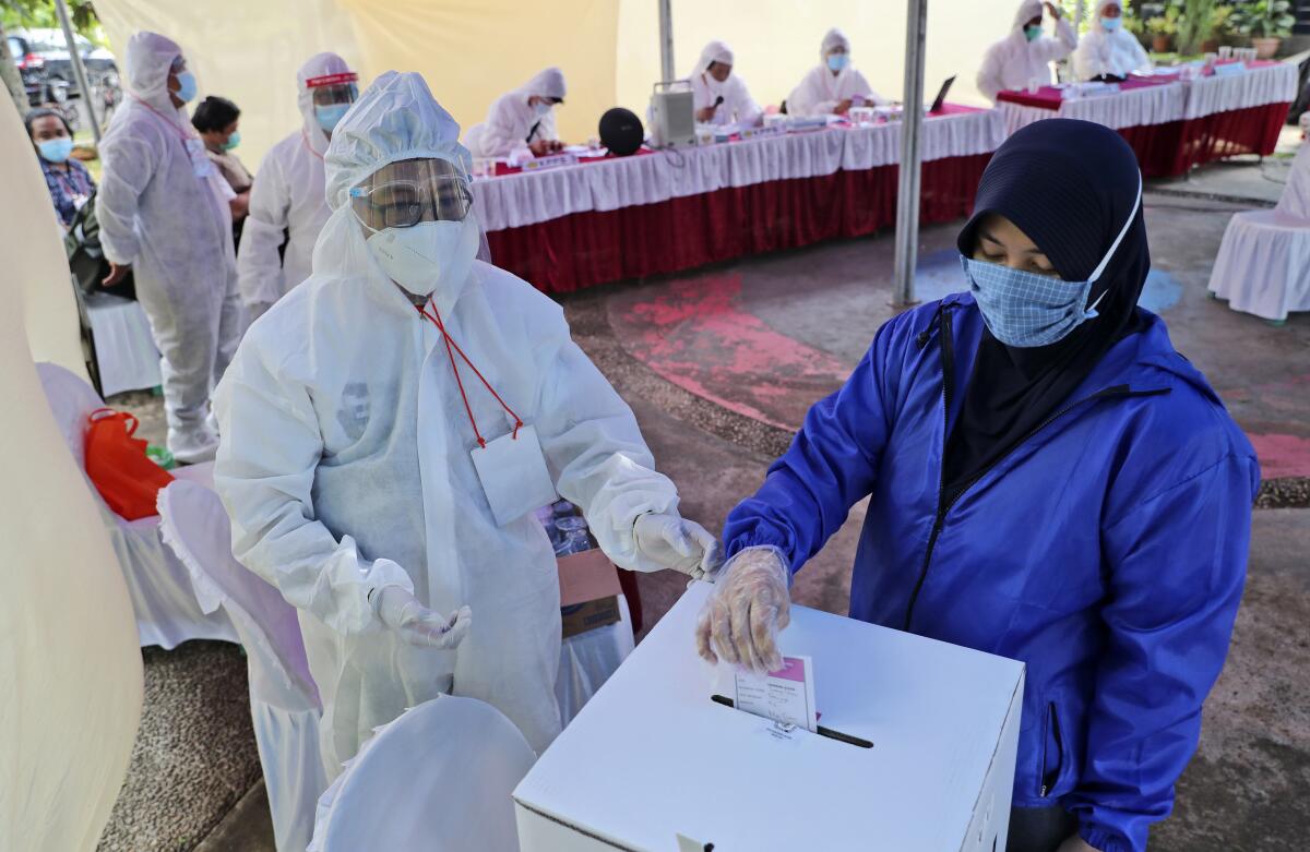 An electoral worker wearing a protective suit as a precaution against the coronavirus outbreak assists a woman to cast her ballot during the regional election at a polling station in Tangerang, Indonesia, Wednesday, Dec. 9, 2020. Indonesia pushed forward with holding previously postponed regional elections on Wednesday amid concerns about the spread of the coronavirus. (AP Photo/Tatan Syuflana)