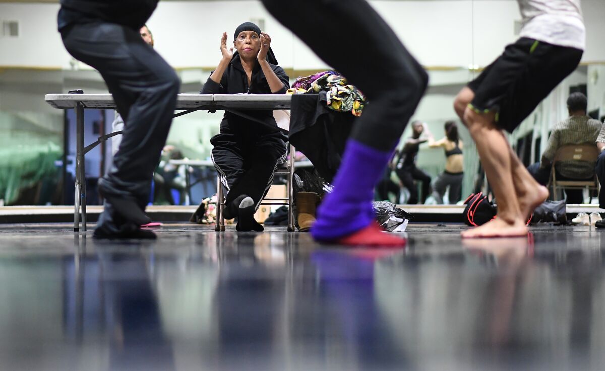 Lula Washington, sitting at a table in mid-clap, watches dancers, whose legs can be seen in the foreground.