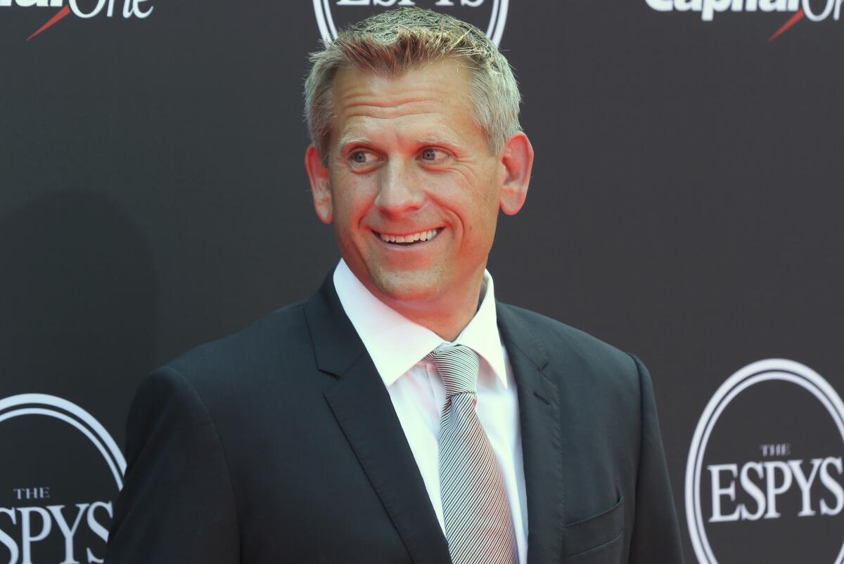 John Brenkus, wearing a dark suit and light gray tie, stands in front of backdrop that reads "The ESPYs" and "Capitol One"