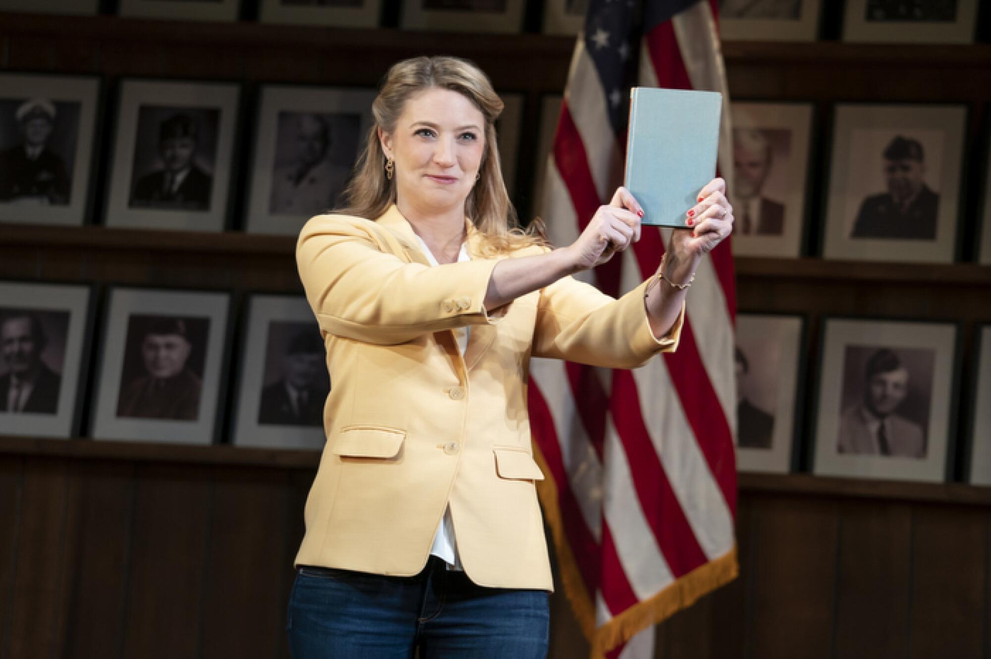 A woman in a yellow blazer and jeans holds up a book 