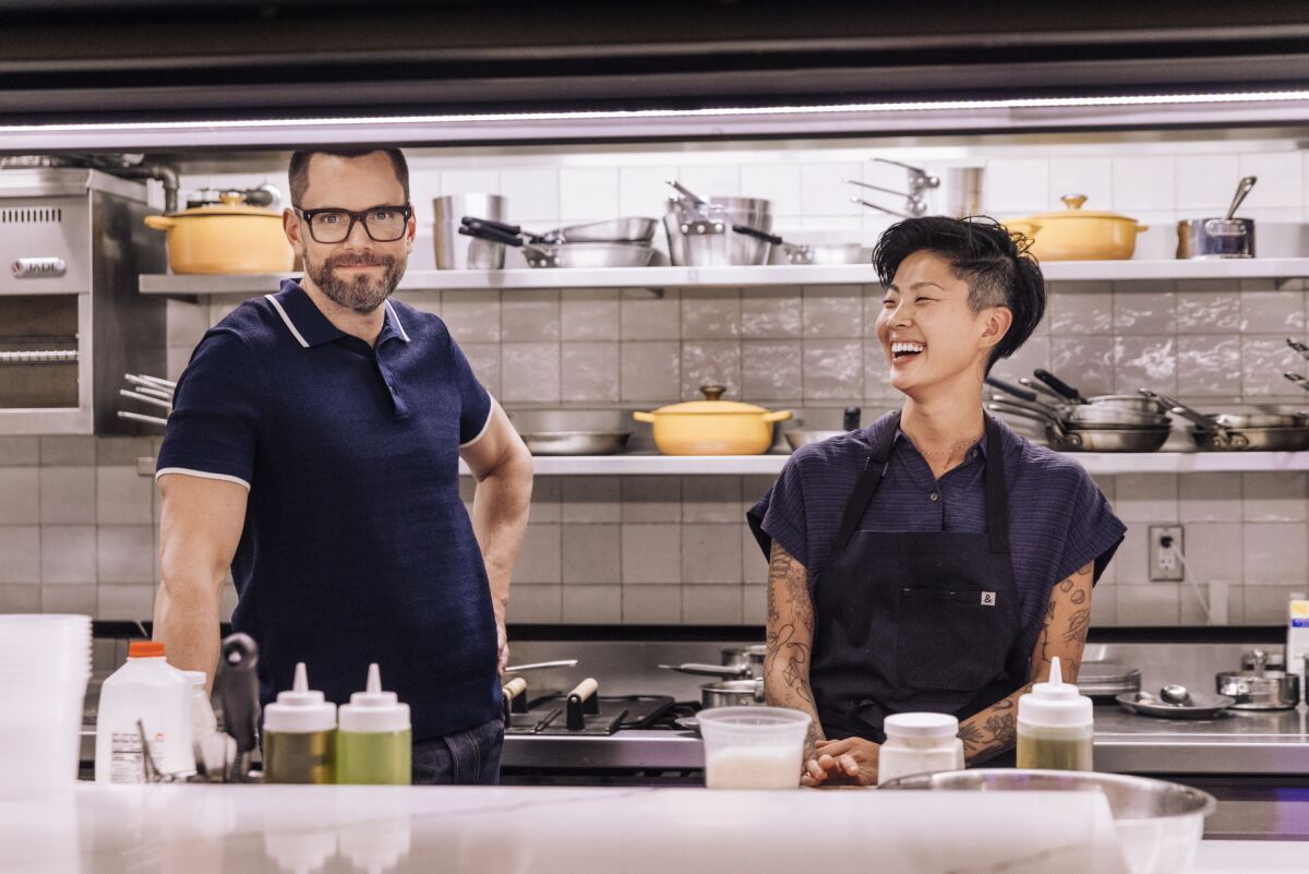 A man smiles at the camera and a woman laughs in a restaurant kitchen.