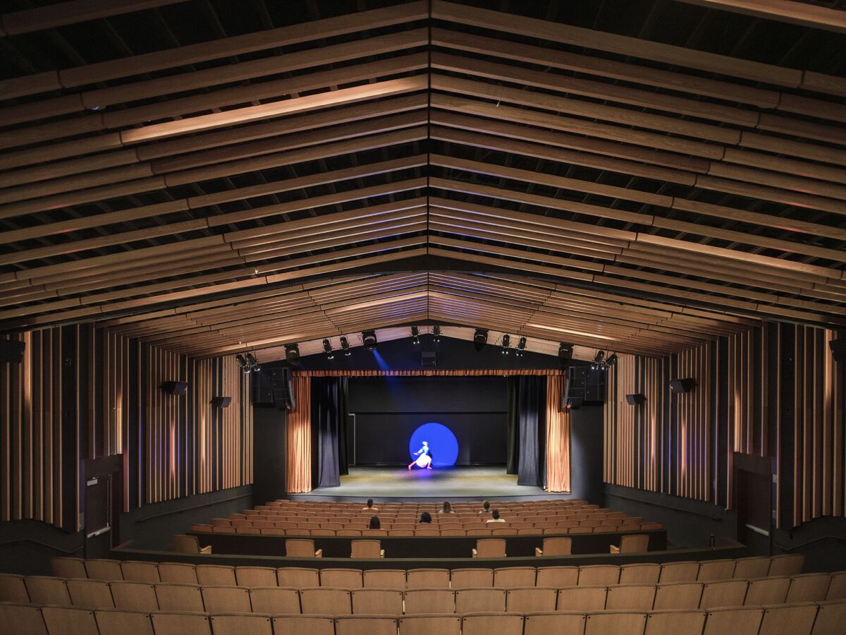 An empty hall is seen with a dancer doing a lunge on stage illuminated by a blue spotlight
