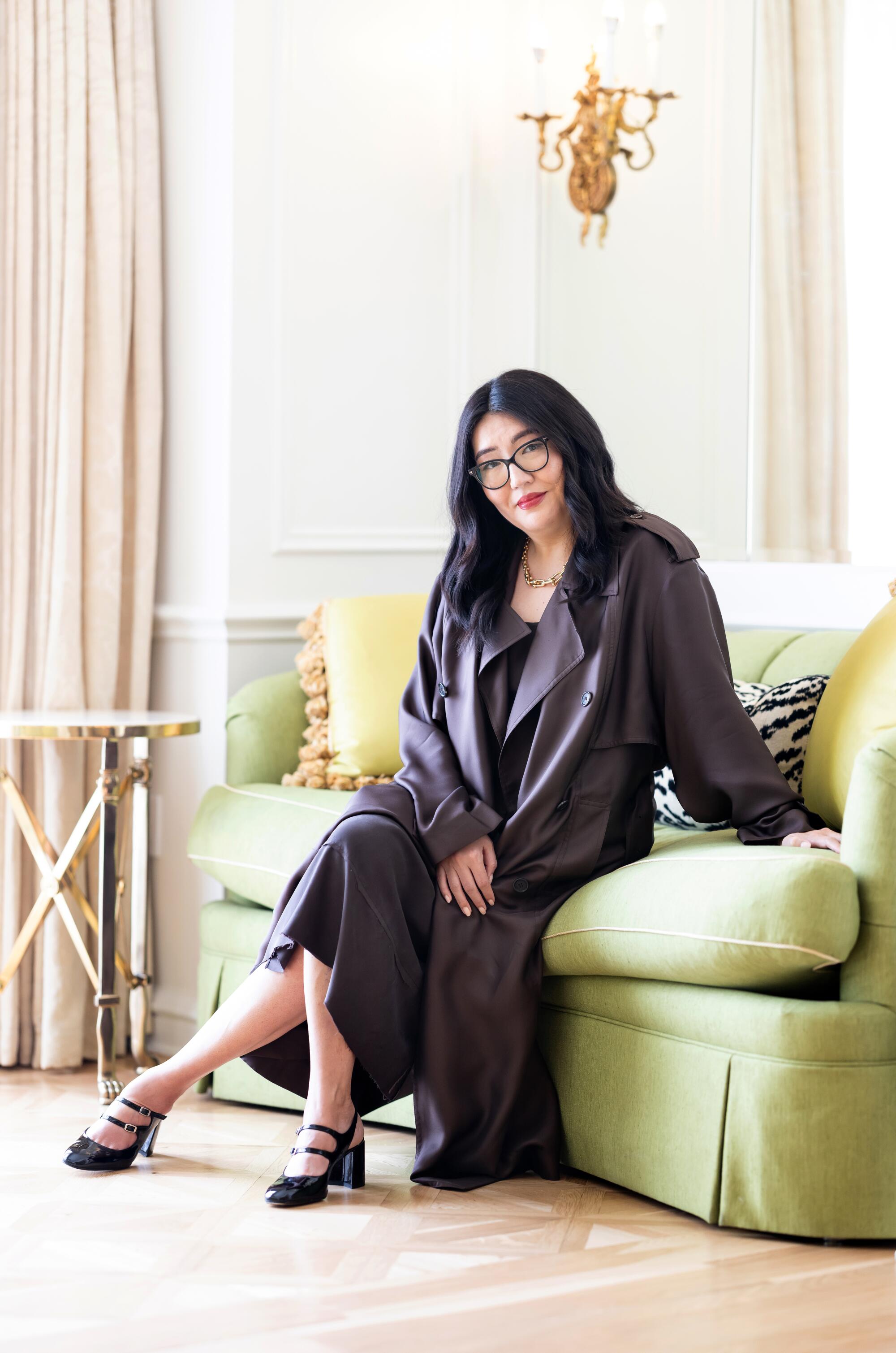 A woman in glasses in a black coat, dress and shoes sits on a bright green couch with her legs crossed at her ankles.