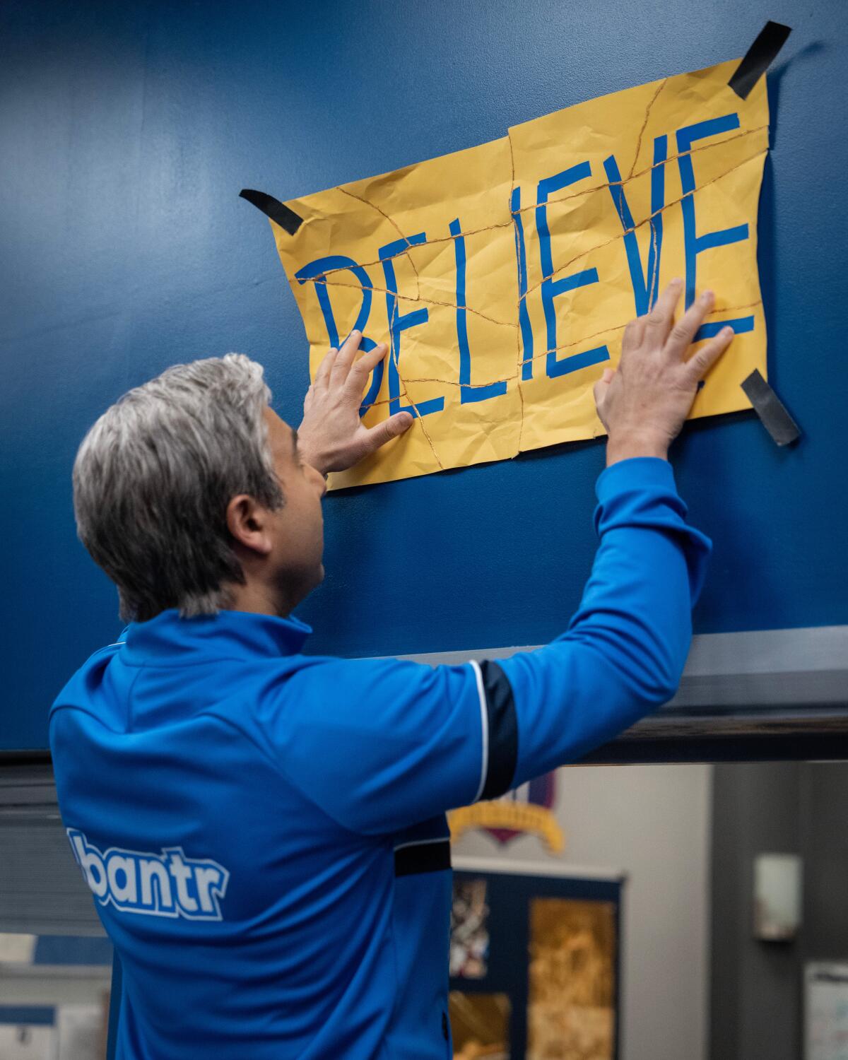 A man hangs a torn and pieced together sign reading "BELIEVE" above a door.
