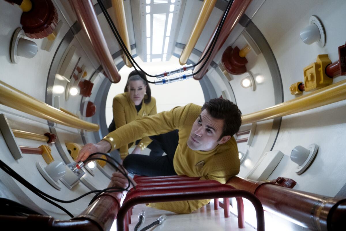 Una and James T. Kirk in yellow and black uniforms, climbing up a red ladder in a narrow tunnel.