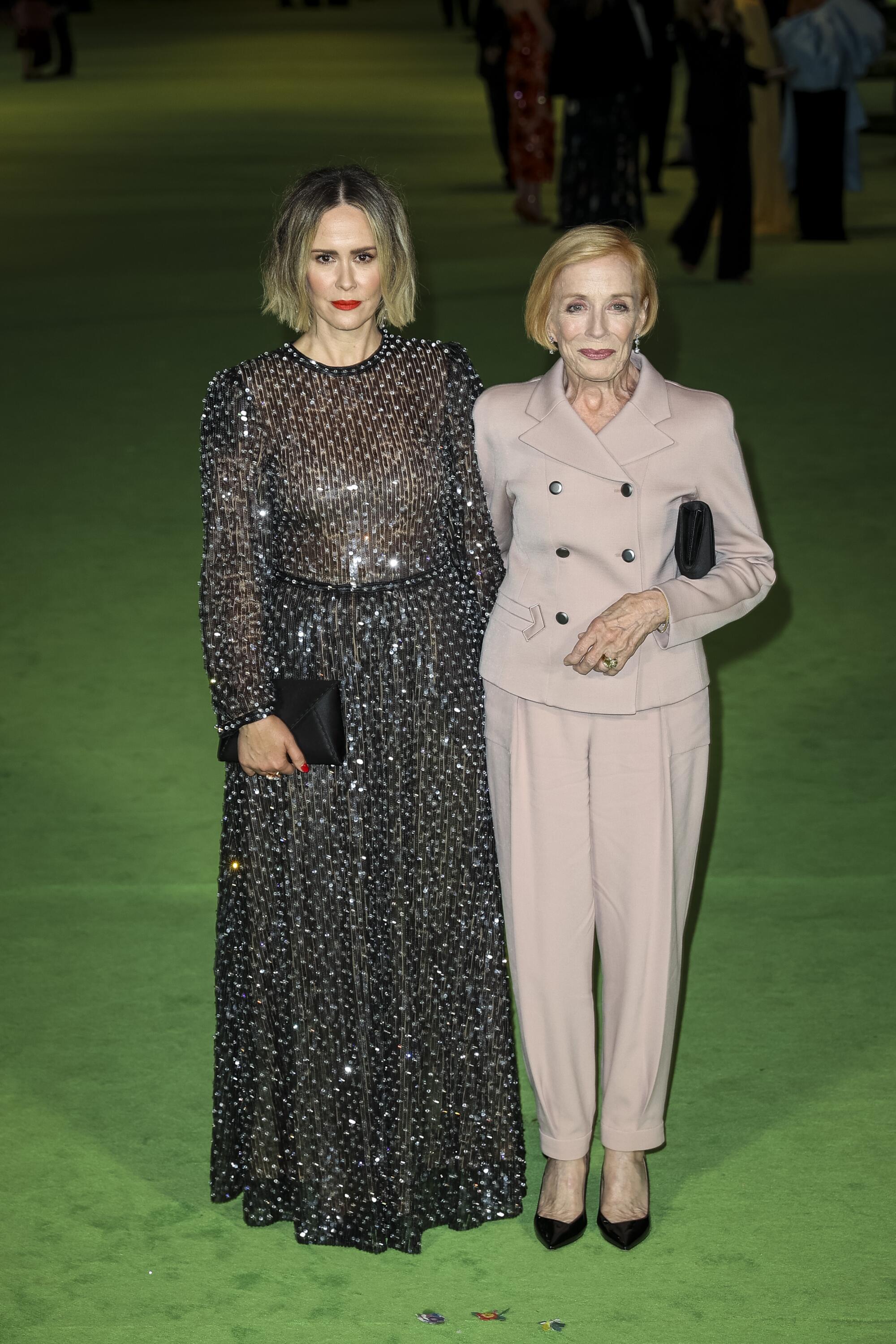 A woman in a black sequined dress next to a woman in a beige pantsuit on the green carpet