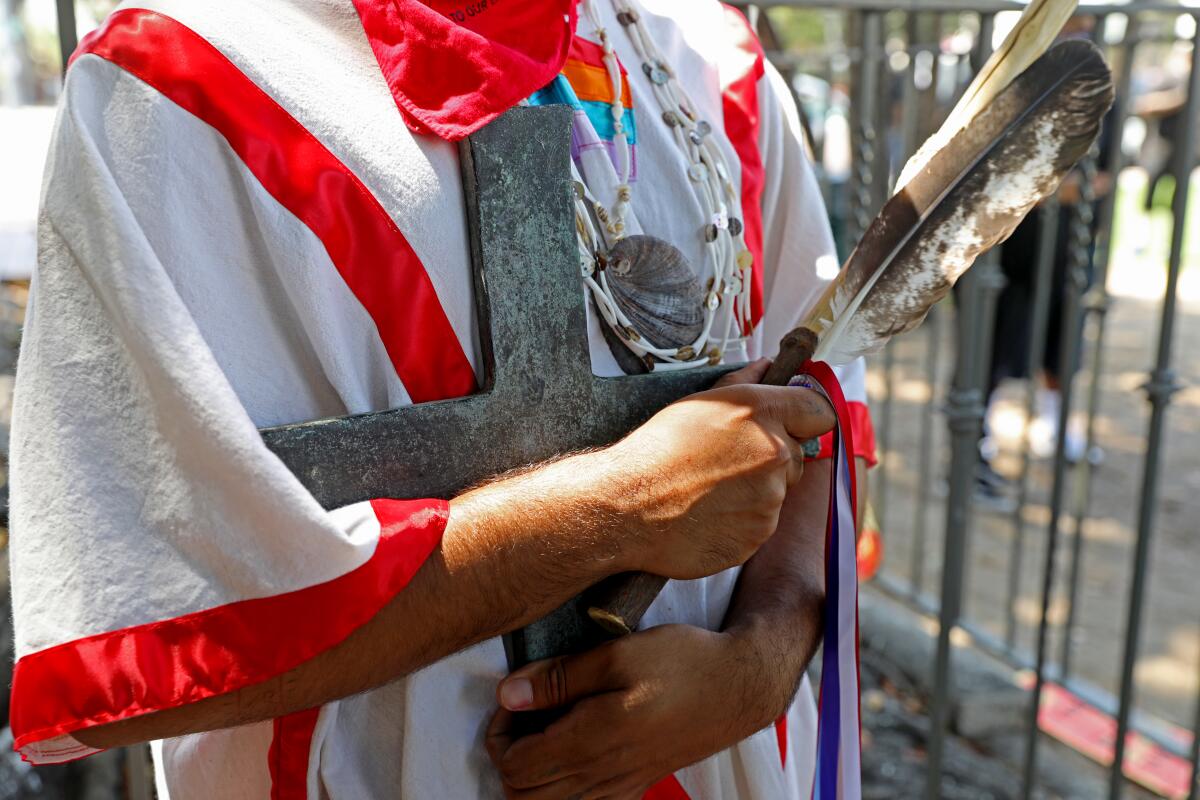 An activist in indigenous clothing holds the cross once held by the statue of Father Junipero Serra 