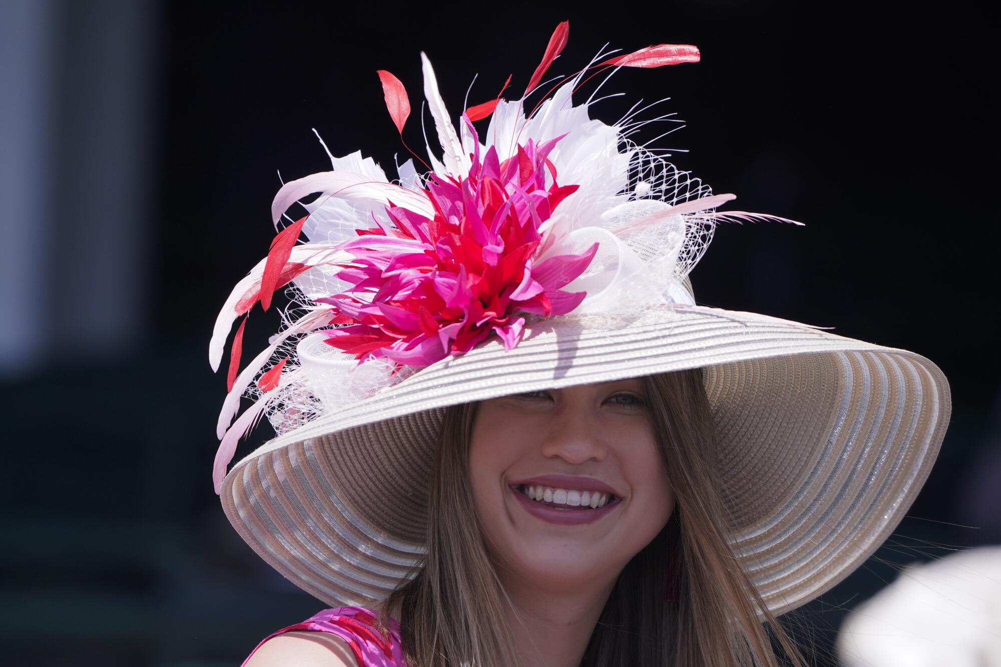 A woman smiles while wearing a white stripped brimmed hat with a pink and red flower and feathers on it.