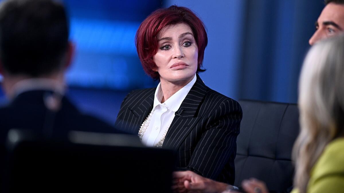 Sharon Osbourne sits on a panel and listens with a serious face in a black pinstripe suit and white shirt
