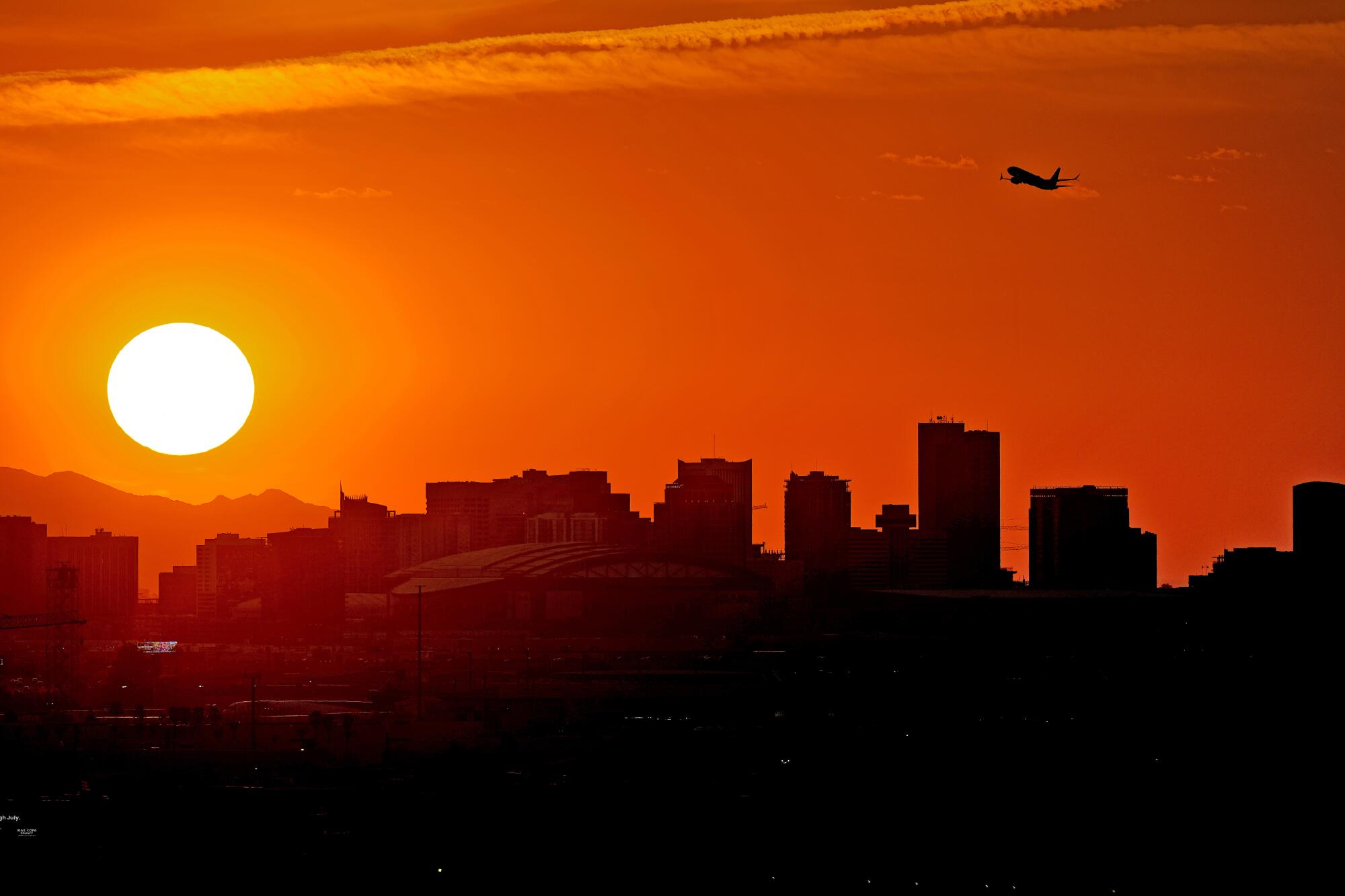 A jet and a city skyline are silhouetted by the sun.