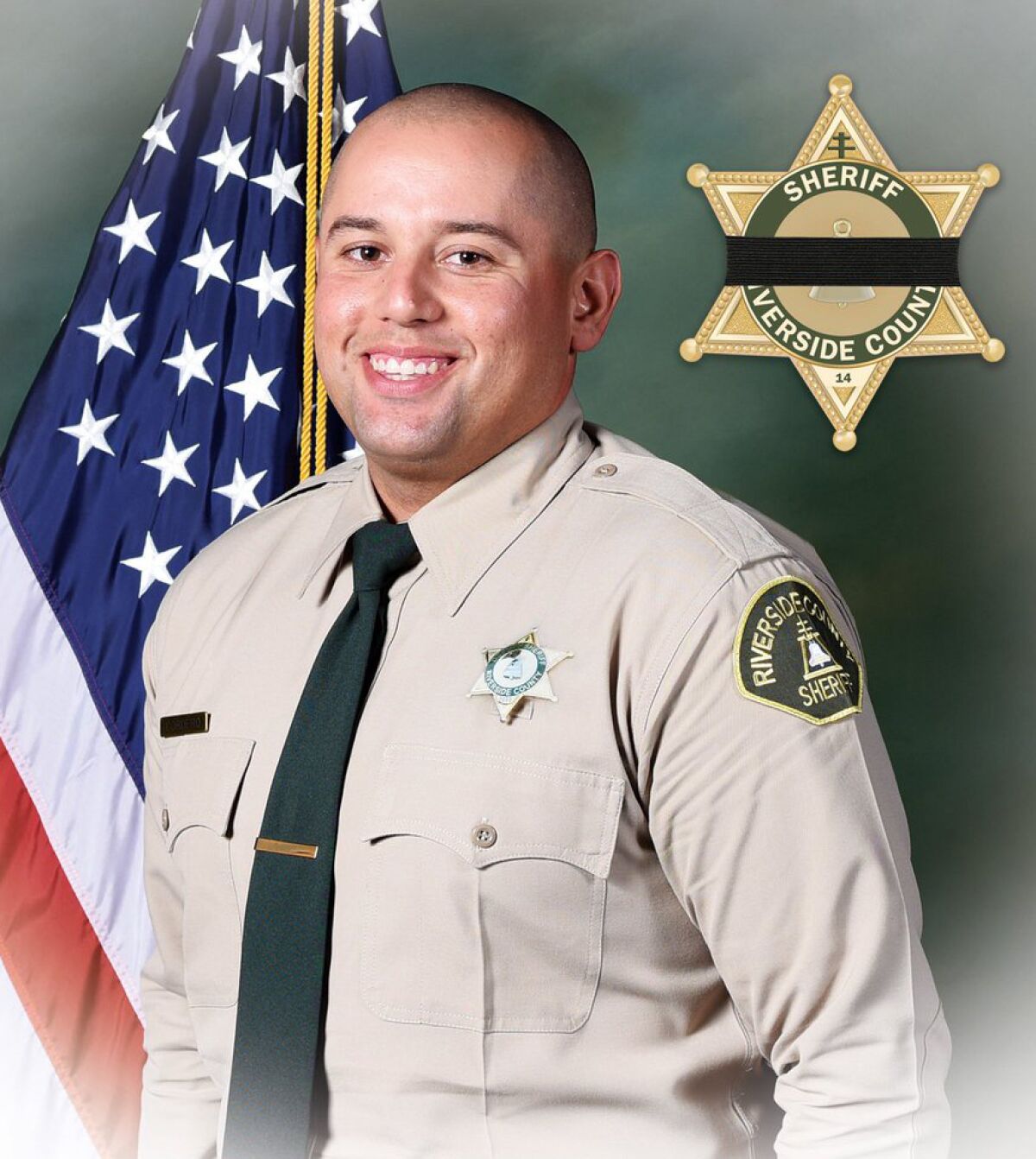 A man in a sheriff's deputy uniform poses for a portrait in front of a U.S. flag and smiles.