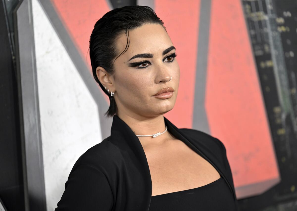 A woman with dark, slicked back hair poses in an all black outfit and a silver choker.