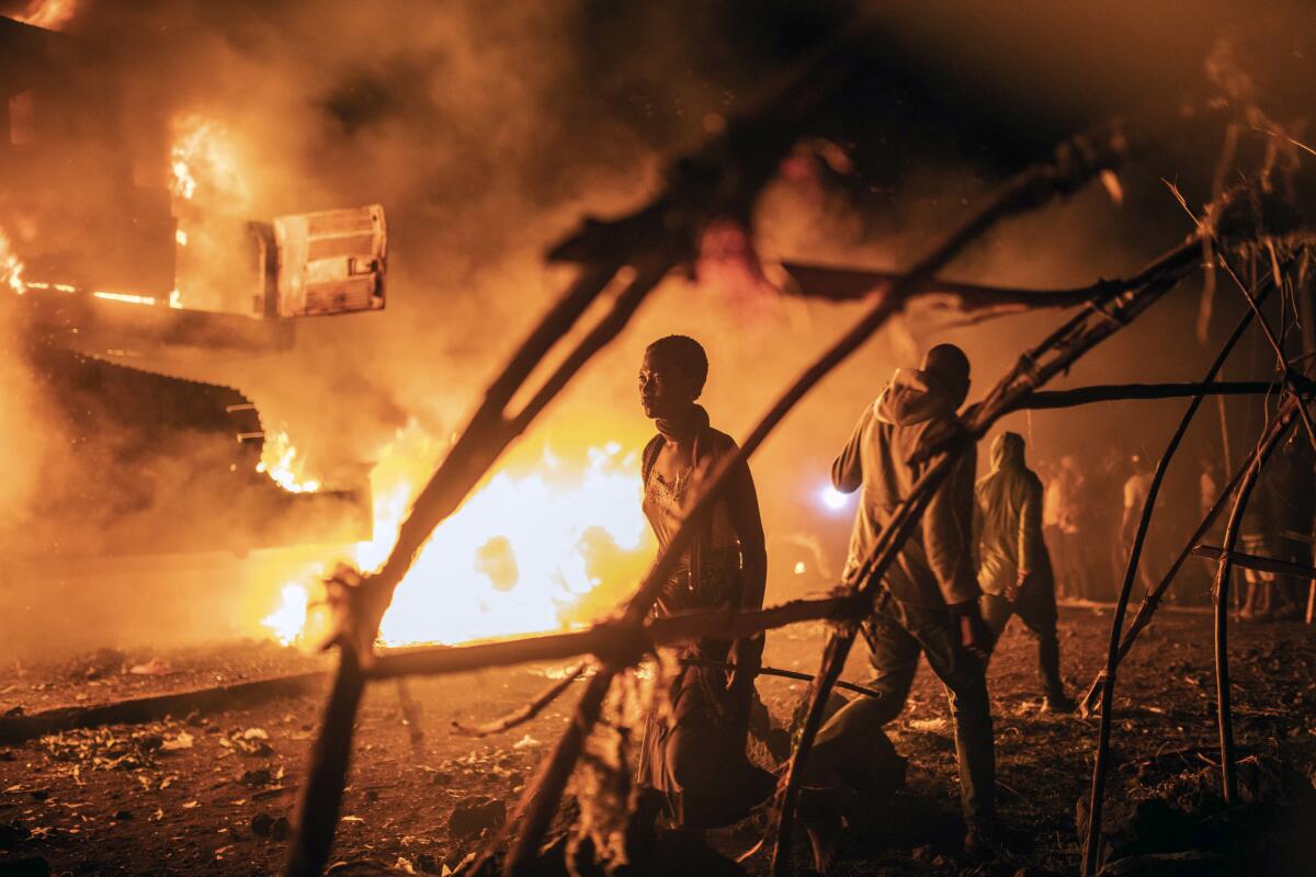 Cars burn in Congo as people walk by.
